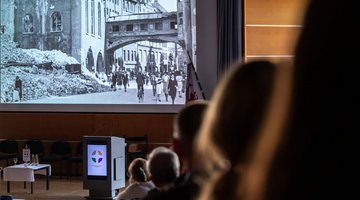 Blick auf eine schwarz-weiße Leinwand, auf die Bilder einer kriegszerstörten Stadt zu sehen sind