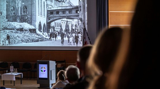 Blick auf eine schwarz-weiße Leinwand, auf die Bilder einer kriegszerstörten Stadt zu sehen sind